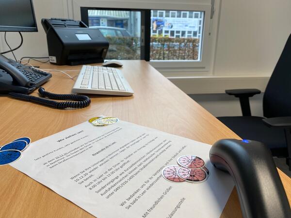 Anträge und Kennzeichen-Plaktten sowie Scanner auf dem Schreibtisch mit Maus und Drucker, dahinter ein großes Fenster welches als Durchreiche dient