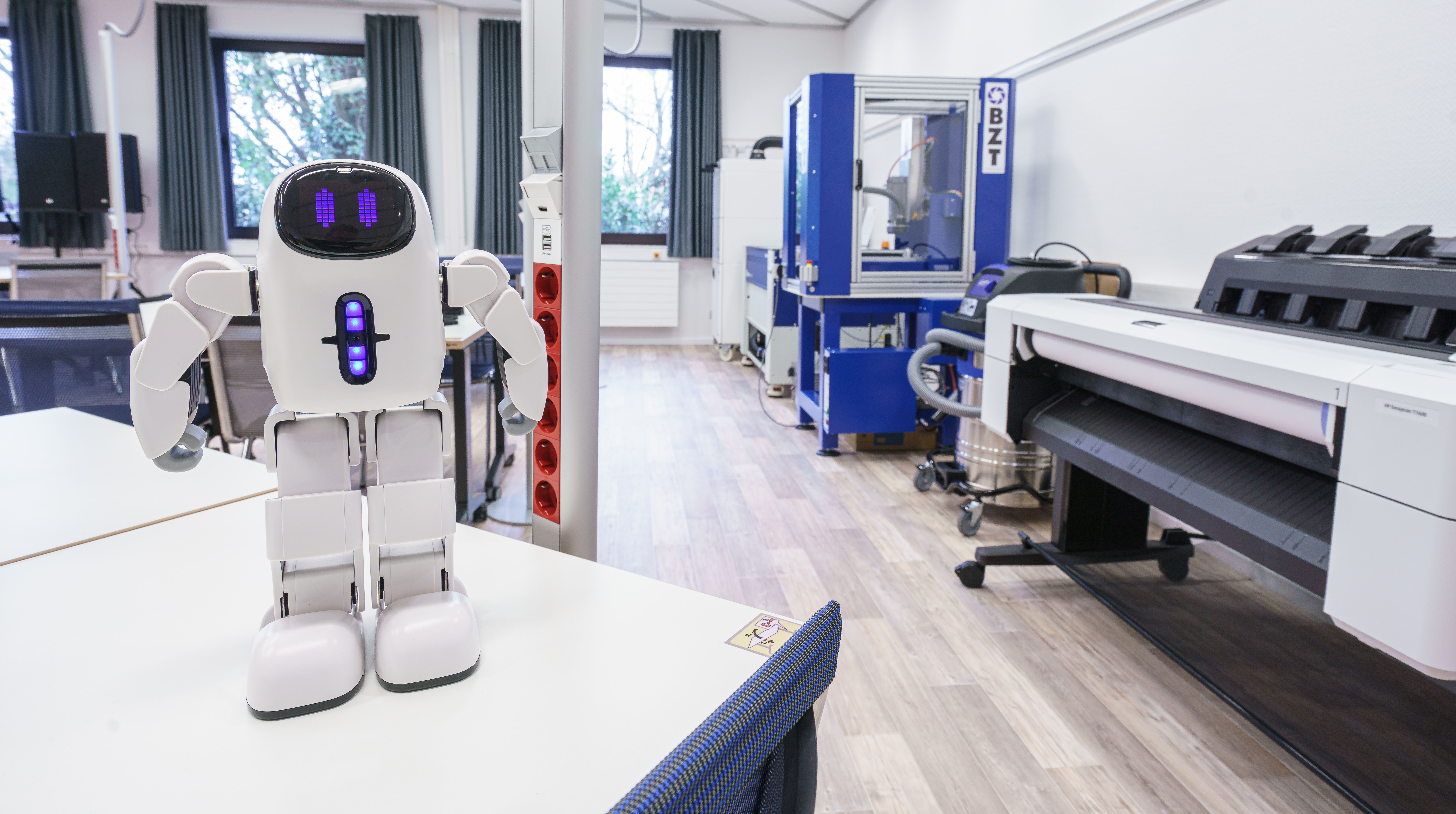 Im Vordergrund ein Roboter auf einem Tisch in einem gro&szlig;em Raum. Im Hintergrund Drucker, Plotter Scanner.