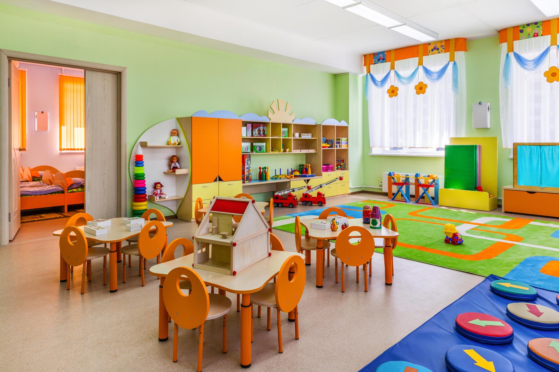 Spielzimmer eines Kindergartens mit grünen Wänden, im Vordergrund drei Tische mit Kinderstühlen.