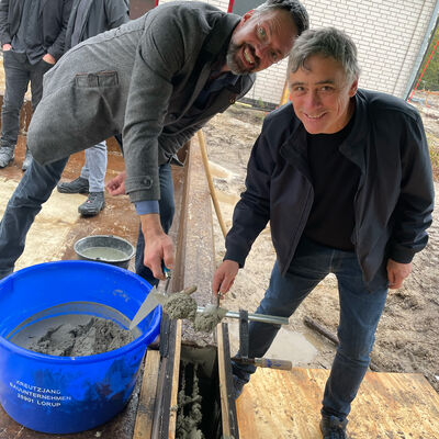 Zwei M&auml;nner beugen sich l&auml;chelnd &uuml;ber eine Baugrube und f&uuml;llen gemeinsam Beton hinein