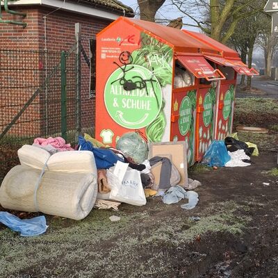 Neben mehreren orangefarbenen Sammelcontainern f&uuml;r Altkleider und Schuhe liegen verschiedene Gegenst&auml;nde auf dem Boden, darunter eine Matratze, Plastikt&uuml;ten, Kleidung und Kartons.