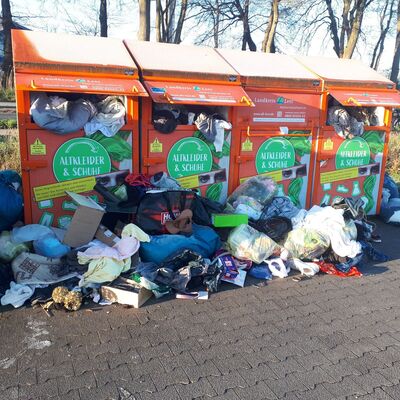 Vor vier orangefarbenen Sammelcontainern f&uuml;r Altkleider und Schuhe liegen zahlreiche Plastikt&uuml;ten, Kartons und Kleidungsst&uuml;cke auf dem Boden. Der Bereich ist stark verm&uuml;llt, die Container sind &uuml;berf&uuml;llt.