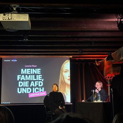 Leonie Plaar sitzt neben Leinwand auf der zu lesen ist "Meine Familie, die AFD und ich". Im Vordergrund sitzt Publikum.