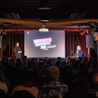 Leonie Plaar steht neben der Leinwand, auf der zu lesen ist "Wer hat's gesagt?". Im Vordergrund sitzt Publikum.