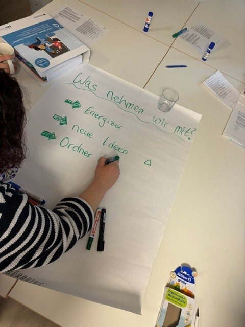 P&auml;dagogin f&uuml;llt ein Papier f&uuml;r eine Flipchart-Tafel aus: "Was nehmen wir mit?" Als Punkte sind schon Energizer, neue Ideen notiert.