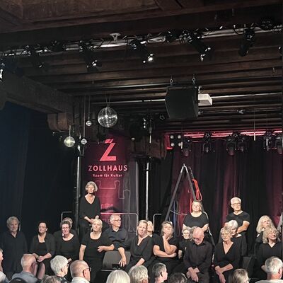 Auff&uuml;hrung von Personen im Zollhaus, Es sind schwarz gekleidete Personen auf der B&uuml;hne zu sehen, im Vordergrund sitzen Zuschauer mit dem Blick zur B&uuml;hne gewandt